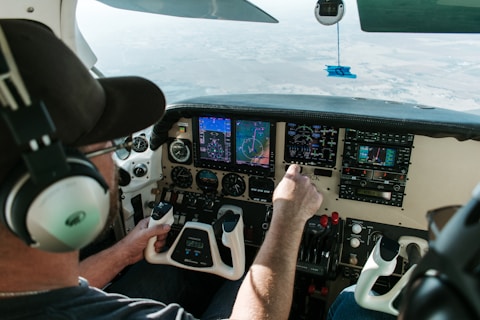 A person wearing a headset is seated in the cockpit of a small aircraft, operating the controls. The instrument panel is active, displaying various digital gauges and navigation screens. The view through the windshield shows a landscape below.