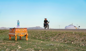 A small, ornately decorated table sits on a grassy field with a plastic bottle and cup on top. In the background, two people ride a motorcycle on a road. A traditional yurt is visible on the right side, set against a vast, open landscape with distant hills under a clear blue sky.