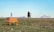 A small, ornately decorated table sits on a grassy field with a plastic bottle and cup on top. In the background, two people ride a motorcycle on a road. A traditional yurt is visible on the right side, set against a vast, open landscape with distant hills under a clear blue sky.