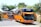 Two large orange tour buses are parked on a paved area surrounded by greenery. The buses have prominent designs on the front and are marked with names like 'Young Guns'. Lush trees and plants can be seen around the area, creating a vivid contrast with the buses.