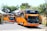 Two large orange tour buses are parked on a paved area surrounded by greenery. The buses have prominent designs on the front and are marked with names like 'Young Guns'. Lush trees and plants can be seen around the area, creating a vivid contrast with the buses.