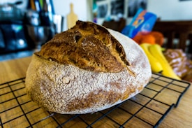 A loaf of crusty artisan bread sits on a cooling rack. The bread has a rough, golden-brown crust with a deep slash across the top. It rests on a wooden countertop with blurred background elements including a cutting board and some colorful fabrics.