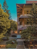 A mid-rise building with multiple balconies and a tiled roof is surrounded by lush greenery, including tall pine trees and dense shrubs. The architecture features wooden accents, and the area appears well-maintained with pathways and a small garden in front.