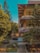 A mid-rise building with multiple balconies and a tiled roof is surrounded by lush greenery, including tall pine trees and dense shrubs. The architecture features wooden accents, and the area appears well-maintained with pathways and a small garden in front.