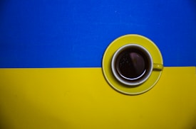 A cup of coffee rests on a yellow saucer placed on a background split into two horizontal halves, blue on top and yellow on the bottom.