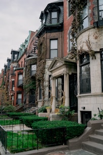 A row of urban townhouse buildings with ornate architectural details such as columns, wrought iron balconies, and large bay windows. The buildings have varying facades of red brick, stone, and ivy-covered walls. The front yards are well-maintained with green bushes, grass, and potted plants. The sky is overcast, adding a slightly gloomy atmosphere.