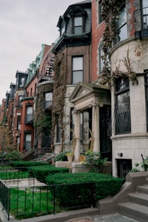 A row of urban townhouse buildings with ornate architectural details such as columns, wrought iron balconies, and large bay windows. The buildings have varying facades of red brick, stone, and ivy-covered walls. The front yards are well-maintained with green bushes, grass, and potted plants. The sky is overcast, adding a slightly gloomy atmosphere.