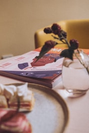 A cozy indoor setting includes a magazine with a colorful illustration on the cover, titled 'Wes Anderson Aesthetics.' It rests on a table next to a blurry detail of dessert on a plate. A small vase with dark red flowers adds a touch of elegance.