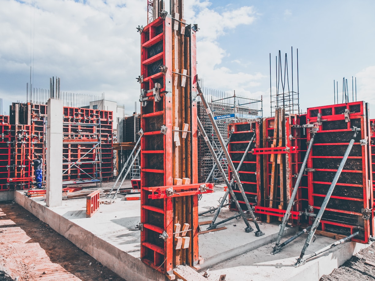 A construction site with multiple concrete formwork structures in place. Metal braces and supports are prominently visible, ensuring the stability of the forms. Concrete foundations and vertical columns are being prepared, with a mix of exposed rebar and newly laid concrete. The overall environment appears organized and active, reflecting ongoing building activities.