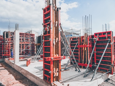 A construction site with multiple concrete formwork structures in place. Metal braces and supports are prominently visible, ensuring the stability of the forms. Concrete foundations and vertical columns are being prepared, with a mix of exposed rebar and newly laid concrete. The overall environment appears organized and active, reflecting ongoing building activities.