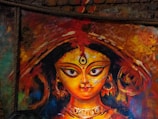 A vibrant and colorful painting of a goddess, featuring large, expressive eyes and intricate details on the headpiece and jewelry. The background is an abstract mix of warm colors, enhancing the divine and vivid appearance of the artwork.