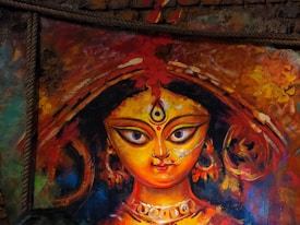 A vibrant and colorful painting of a goddess, featuring large, expressive eyes and intricate details on the headpiece and jewelry. The background is an abstract mix of warm colors, enhancing the divine and vivid appearance of the artwork.