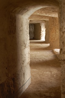 A dimly lit passageway featuring textured stone walls and a dusty floor, curving gently to the right. Arched entrances and natural light filtering in create a mysterious ambiance.