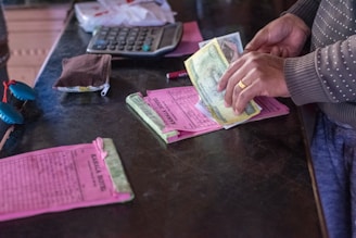 A person is counting paper money while standing at a counter. Various objects are present including a calculator, a small pouch, and papers with printed text. The person's hands are visible, and they are wearing a polka-dotted sweater.
