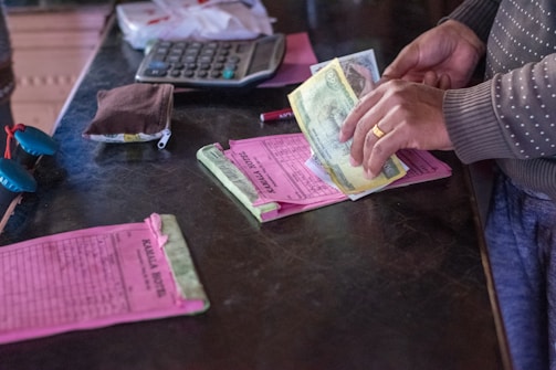 A person is counting paper money while standing at a counter. Various objects are present including a calculator, a small pouch, and papers with printed text. The person's hands are visible, and they are wearing a polka-dotted sweater.