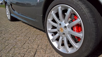 A close-up view of a silver alloy wheel with a visible red brake caliper, attached to a dark-colored car parked on a cobblestone surface. The tire brand is visible on the sidewall, and the MG emblem is centered on the wheel.