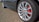 A close-up view of a silver alloy wheel with a visible red brake caliper, attached to a dark-colored car parked on a cobblestone surface. The tire brand is visible on the sidewall, and the MG emblem is centered on the wheel.