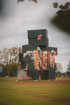 A structure made of stacked shipping containers painted in dark colors, featuring large mural art of human figures and abstract elements. The artwork includes depictions of people and a sliced fruit motif on the upper section. The building is situated in a grassy area with trees in the background under an overcast sky.