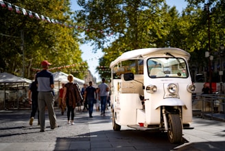 A white tuk-tuk is parked on a paved pedestrian street. Several people are walking in the background under strings of colorful pennant flags. The street is lined with trees that provide shade, and the atmosphere suggests a casual, sunny day.