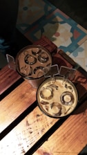 Two cold drinks in clear plastic cups, filled with ice cubes, are placed on a wooden surface. The drinks appear to be a type of iced coffee. The surface they are on has a dark, natural wood grain, and there is decorative tile flooring visible in the background with floral patterns in blue, yellow, and red tones.