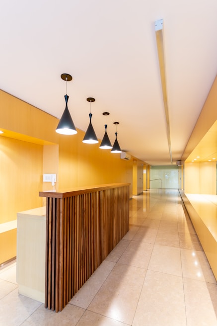 A modern interior space with a long hallway and sleek design. Wooden paneling forms a partition with overhead pendant lights casting a warm glow. The walls and ceiling feature a light wood finish, enhancing the minimalist aesthetic. The floor is made of polished tiles, contributing to the clean and contemporary look.