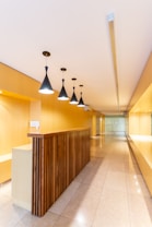 A modern interior space with a long hallway and sleek design. Wooden paneling forms a partition with overhead pendant lights casting a warm glow. The walls and ceiling feature a light wood finish, enhancing the minimalist aesthetic. The floor is made of polished tiles, contributing to the clean and contemporary look.