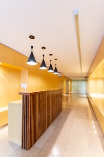 A modern interior space with a long hallway and sleek design. Wooden paneling forms a partition with overhead pendant lights casting a warm glow. The walls and ceiling feature a light wood finish, enhancing the minimalist aesthetic. The floor is made of polished tiles, contributing to the clean and contemporary look.