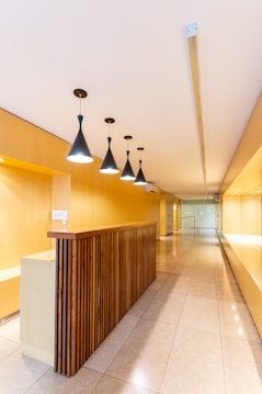 A modern interior space with a long hallway and sleek design. Wooden paneling forms a partition with overhead pendant lights casting a warm glow. The walls and ceiling feature a light wood finish, enhancing the minimalist aesthetic. The floor is made of polished tiles, contributing to the clean and contemporary look.