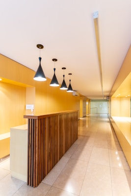 A modern interior space with a long hallway and sleek design. Wooden paneling forms a partition with overhead pendant lights casting a warm glow. The walls and ceiling feature a light wood finish, enhancing the minimalist aesthetic. The floor is made of polished tiles, contributing to the clean and contemporary look.