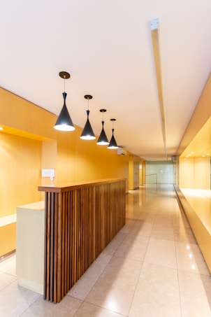 A modern interior space with a long hallway and sleek design. Wooden paneling forms a partition with overhead pendant lights casting a warm glow. The walls and ceiling feature a light wood finish, enhancing the minimalist aesthetic. The floor is made of polished tiles, contributing to the clean and contemporary look.
