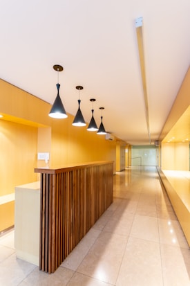 A modern interior space with a long hallway and sleek design. Wooden paneling forms a partition with overhead pendant lights casting a warm glow. The walls and ceiling feature a light wood finish, enhancing the minimalist aesthetic. The floor is made of polished tiles, contributing to the clean and contemporary look.