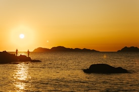 A breathtaking sunset over a calm sea with a clear sky, casting golden hues across the water. Silhouettes of two people stand on rocky formations, adding depth to the serene coastal landscape. Distant islands are visible on the horizon, enhancing the scenic beauty.