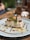 A dessert with a creamy texture topped with a slice of orange and a mint leaf sits on a light blue plate. The dessert is garnished with crumbled toppings and drizzled with caramel sauce. The background includes a blurred scene of a table setting with glasses and vaguely defined objects.