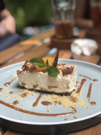 A dessert with a creamy texture topped with a slice of orange and a mint leaf sits on a light blue plate. The dessert is garnished with crumbled toppings and drizzled with caramel sauce. The background includes a blurred scene of a table setting with glasses and vaguely defined objects.