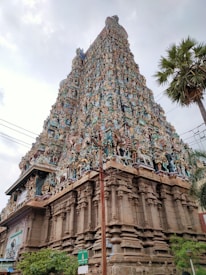 A large, intricately decorated temple tower with detailed carvings and sculptures in a variety of colors. The structure is imposing and prominently features numerous figures and ornate designs, characteristic of traditional South Indian architecture. A few palm trees and some electrical wires are visible near the temple, with a cloudy sky in the background.