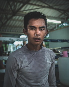 A young man with short hair stands in a gym setting, wearing a gray, long-sleeve athletic shirt. His skin glistens with sweat, indicating recent physical exertion. The background shows gym equipment and a partially visible boxing ring, all under a metal roof structure.