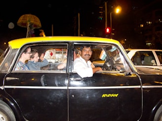 A man driving a black and yellow taxi is smiling at the camera. Two passengers are seated in the back, engaged in conversation. The taxi is surrounded by traffic at night, and an Indian flag is displayed on the dashboard.