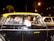 A man driving a black and yellow taxi is smiling at the camera. Two passengers are seated in the back, engaged in conversation. The taxi is surrounded by traffic at night, and an Indian flag is displayed on the dashboard.