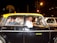 A man driving a black and yellow taxi is smiling at the camera. Two passengers are seated in the back, engaged in conversation. The taxi is surrounded by traffic at night, and an Indian flag is displayed on the dashboard.