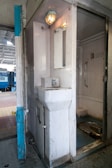 A compact train lavatory with basic amenities, including a small sink, a mirror, and a metal squat toilet. The walls and fixtures are white and slightly worn, reflecting utilitarian design. A light fixture is mounted above the mirror, providing illumination.
