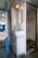 A compact train lavatory with basic amenities, including a small sink, a mirror, and a metal squat toilet. The walls and fixtures are white and slightly worn, reflecting utilitarian design. A light fixture is mounted above the mirror, providing illumination.