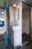 A compact train lavatory with basic amenities, including a small sink, a mirror, and a metal squat toilet. The walls and fixtures are white and slightly worn, reflecting utilitarian design. A light fixture is mounted above the mirror, providing illumination.