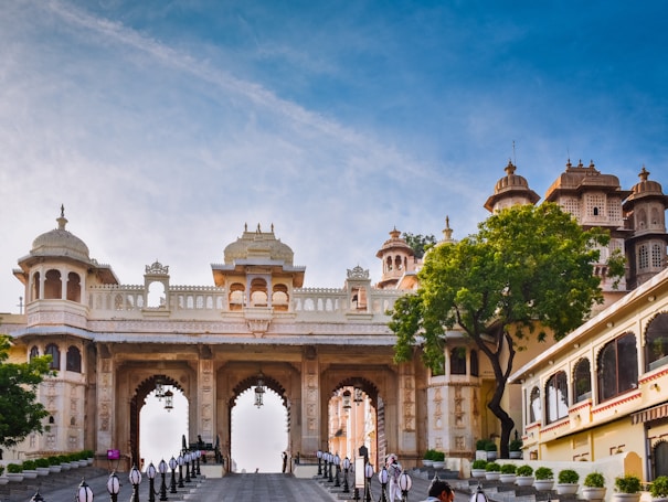 A grand architectural structure featuring ornate arches and decorative elements, with a blend of traditional and historical design. The building is adorned with detailed carvings and has a large tree to the right. The sky is clear, providing a serene setting.