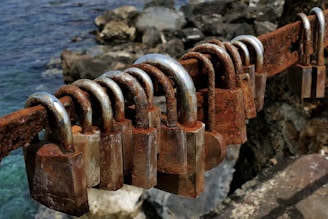 Several rusty padlocks are attached to a decaying metal bar, with a rocky shoreline and the sea visible in the background.