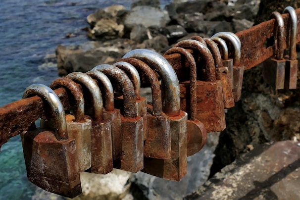 Several rusty padlocks are attached to a decaying metal bar, with a rocky shoreline and the sea visible in the background.