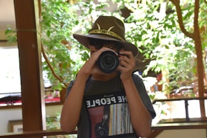 A person wearing a brown cowboy hat is holding a camera in front of their face, capturing a photograph. They are in an indoor space filled with lush green plants and natural light. The person is wearing a dark T-shirt.