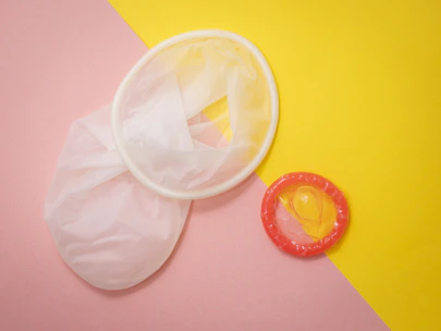 Two contraceptives, a female condom and a male condom, are placed on a pastel pink and bright yellow background. The female condom is white and translucent, while the male condom is transparent with a red ring.
