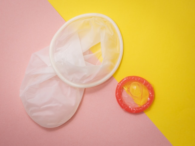 Two contraceptives, a female condom and a male condom, are placed on a pastel pink and bright yellow background. The female condom is white and translucent, while the male condom is transparent with a red ring.