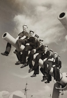 A group of sailors in uniform is sitting on a large naval gun aboard a ship. The scene is set against a backdrop of a partly cloudy sky. The sailors appear to be relaxed and smiling.