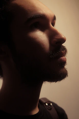 A restrained editorial-style image of Profit Elijah seated contemplatively with shadows playing across his face and a textured backdrop.